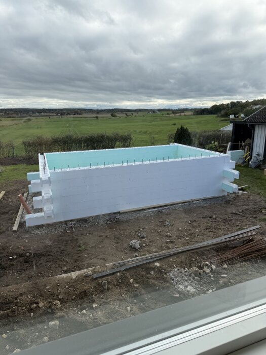 Byggarbetsplats med en halvfärdig pool av vita block, inre förstärkningar och en öppen gräsyta i bakgrunden under molnig himmel.