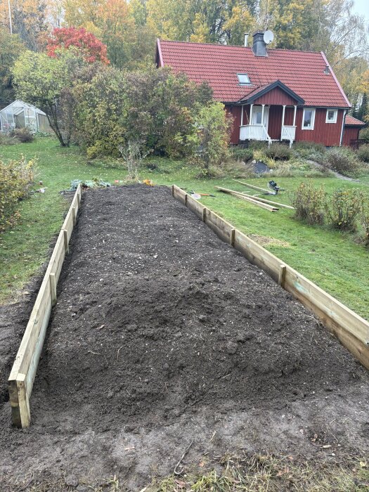 Byggd träram runt odlingsland i trädgård framför röd stuga med rött tak och omgivande grönska.