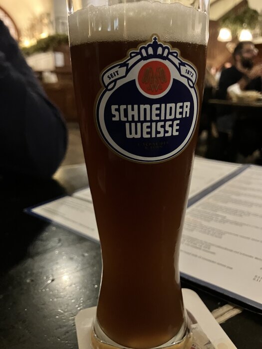 Ett glas Schneider Weisse öl på bordet, märkt med logotypen, i en restaurangmiljö.