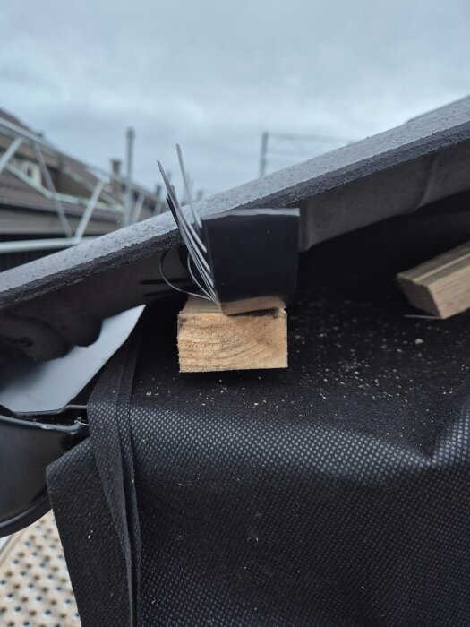 Bit av trä under takpanna med svart byggplast och metallkrokar ovanpå, i bakgrunden syns delar av ett hustak och himmel.