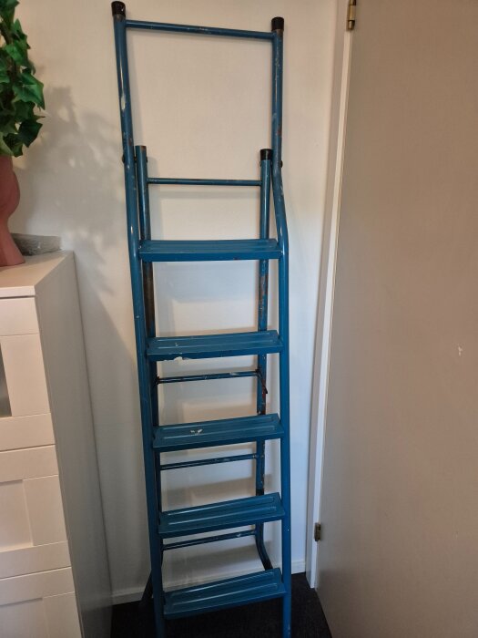 Blå metallstege med runda steg, hopvikt mot en vägg.
