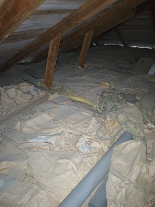 Vindsutrymme med synliga träbjälkar, isoleringsmaterial och plast- eller pappersskydd på golvet under ett plåttak. Ingen mekanisk ventilation syns.