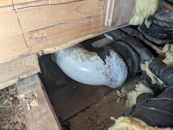 Plastavloppsrör och gjutjärnsrör i en trång utrymme under träskiva, reparerat med grå massa och rubber seal för att stoppa läckage.