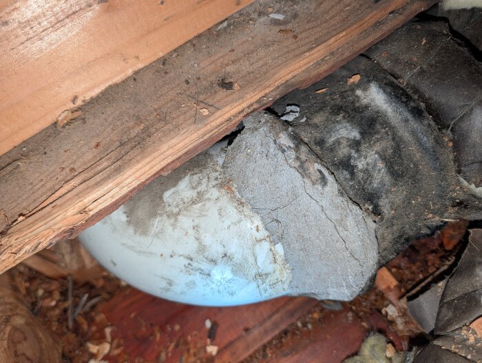 Skadat avloppsrör med synlig skarv mellan plast- och gjutjärnsdel, omgiven av trä och grå tätmassa som delvis har lossnat.