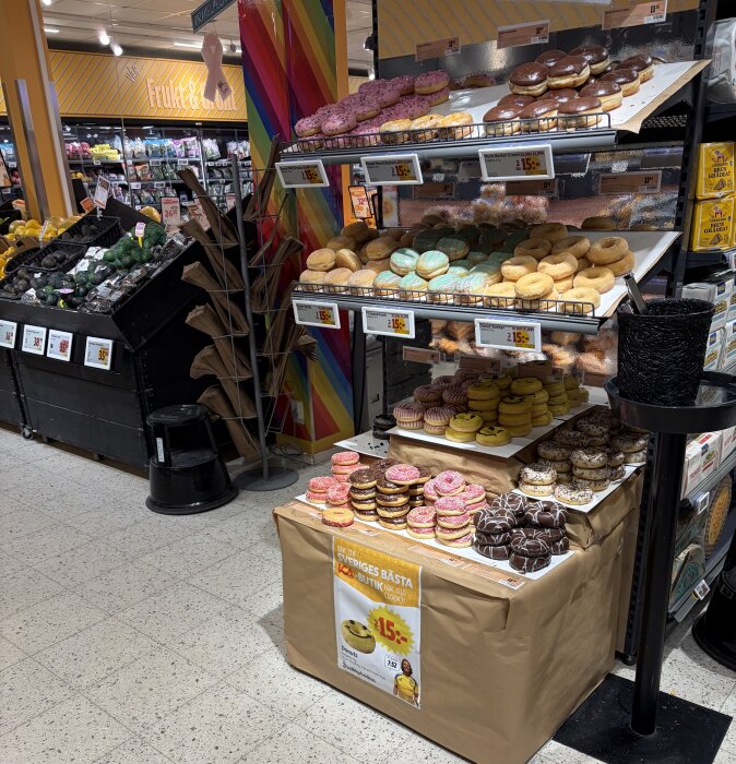 Hylla med olika sorters munkar i en mataffär, inklusive choklad, rosa glasyr och ringlade mönster, med frukt- och grönsaksavdelning i bakgrunden.