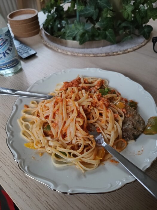 Pasta med tomatsås och grönsaker på tallrik, bredvid Dafgårds italienska köttfärsbiff, på ett träbord med en flaska vatten och växt i bakgrunden.