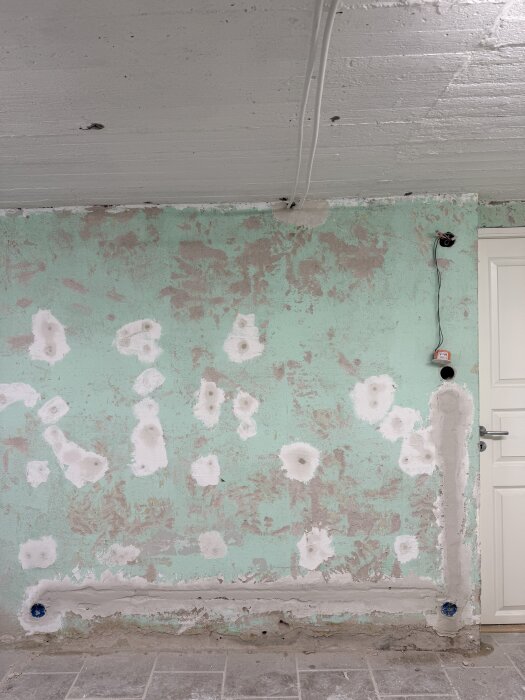 En grönt målad vägg med spacklade områden och eldragningar som har täckts, med ett hål för en eluttag synligt. Väggen är under renovering.
