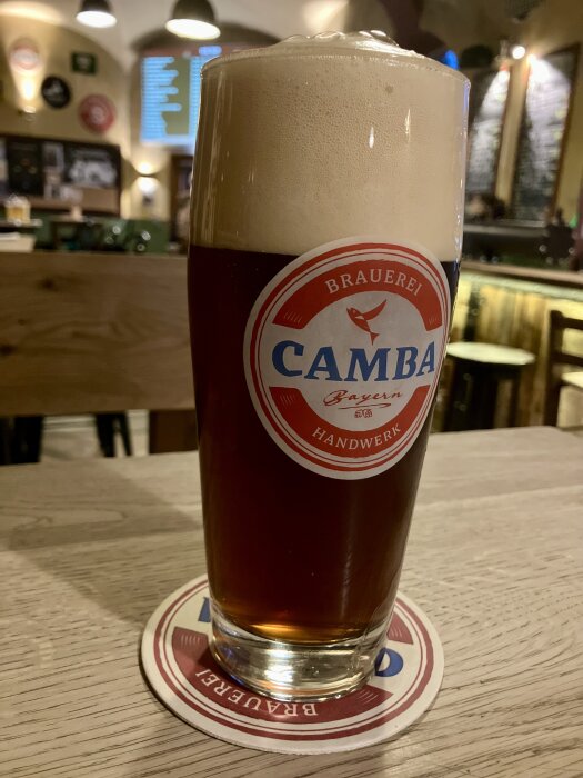 Glas med mörkt öl och skumhuvud, placerat på ett underlägg med texten "CAMBA" i en bar- eller pubmiljö.
