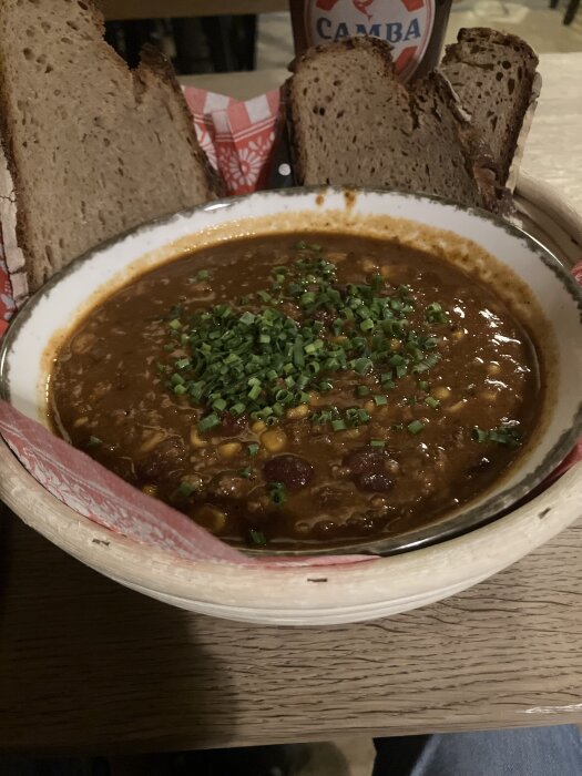Skål med chili con carne toppad med gräslök, omgiven av brödbitar på en träbänk. En del av en flaska syns i bakgrunden.