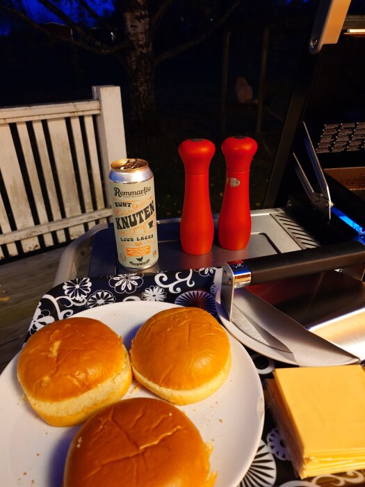 Hamburgerbröd på en tallrik, en ölburk, två röda kryddkvarnar, och ostskivor bredvid en grill på en kvällsbelyst altan.