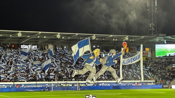 Fotbollsfans med blåvita flaggor och stor banderoll med texten "Allsvenskan" på läktaren under kvällsmatch på arenan.