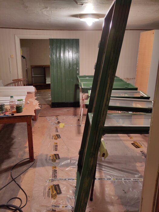 Grönt målat snickeri står lutat mot ett bord i ett rum under renovering, med plast på golvet och en taklampa i bakgrunden.