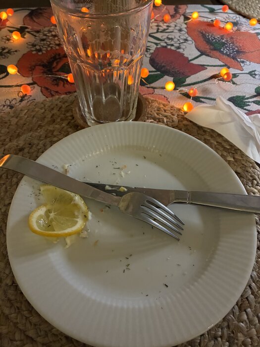 Tom tallrik med citronskivor och bestick, glas i bakgrunden. Bord med blommig duk och orange belysning.