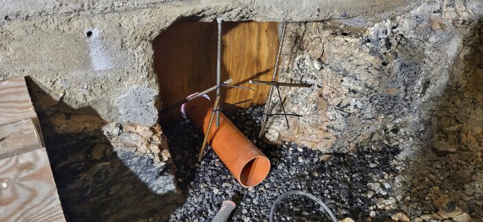 Orange dräneringsrör genom berg med armeringsjärn och grovt grus; del av bygge där berg och betong möts.