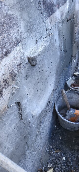 Betongvägg med synliga sprickor och fästen för armering, en säkerhetsbalk och en plasthink fyllt med verktyg och material vid sidan.