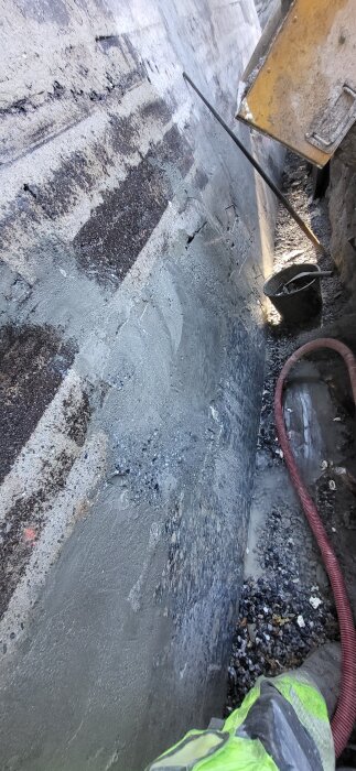 Betongvägg med nyligen tillsatt isolering och armering, röd slang och en arbetsbyxa synlig i bild. Grus ligger längs botten av väggen.