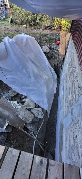 Markarbete vid husvägg med grus, trä, isolering och plastduk. Väggen visar tecken på slammning och borrning för vatten och el.