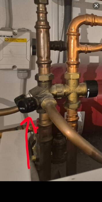 Påfyllningsventil för värmesystem med pilar som visar riktning, omgiven av kopparrör och en säkerhetsbrytare i bakgrunden.
