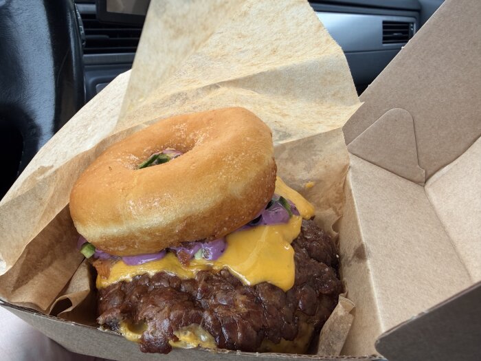 Hamburgare med en munk som överbröd, smält ost och rödlök i en pappersbehållare.