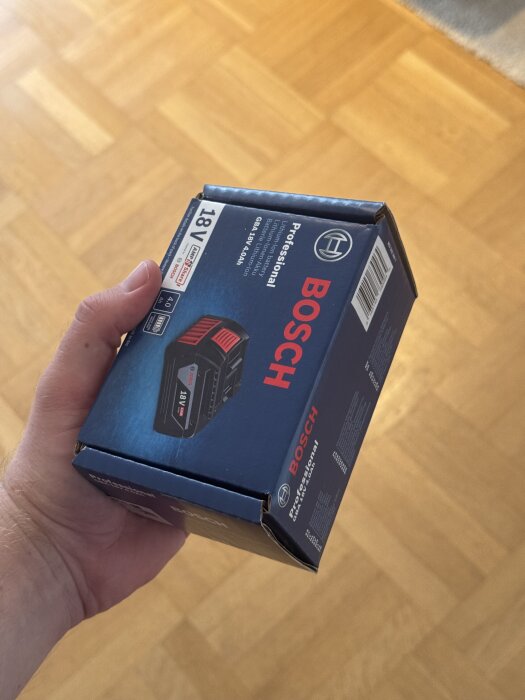 En person håller i en förpackning med ett Bosch 18V batteri på ett trägolv.