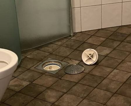 Golvbrunn i badrum med insats och galler urtagna och ligger bredvid på klinkergolv. Toalett synlig i vänstra delen av bilden.