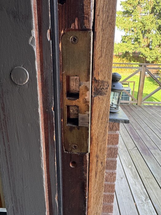 Gammalt dörrlås i träkarm, behöver renovering för att eventuellt kunna montera ett Yale-lås, uteplats och grönska syns i bakgrunden.
