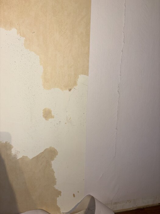 Vägg i vardagsrum med borttagen tapet delvis avtäckt, blottar yta med tecken på möjligen blåbetong.