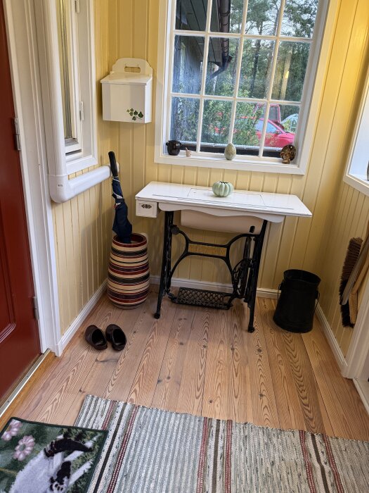 Ljust trägolv efter såpskurning, röda dörren, fönster med utsikt, vit hallmöbel med svart underrede, randig matta, och paraplyställ.