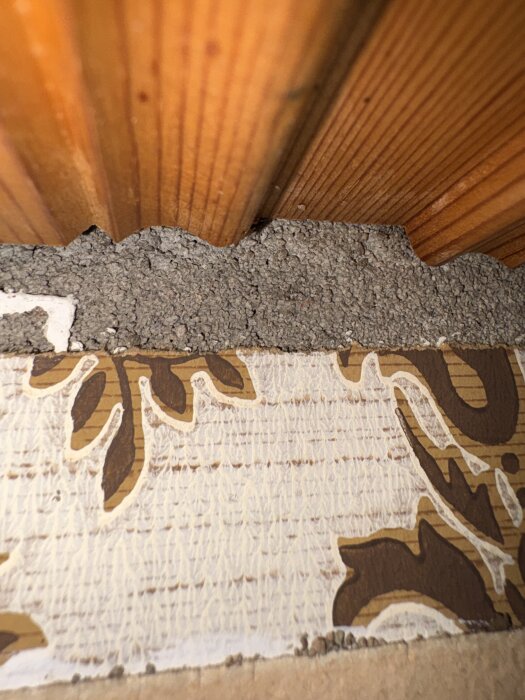 Närbild av en väggdetalj med träpanel ovanpå tapet med brunt bladmönster, synlig övergång mellan de två materialen.