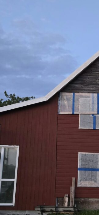 Rött hus med ny vindskiveplåt på taket, gamla och nya sektioner sammansmälter vid vinkeln, blå tejp på fönstren.