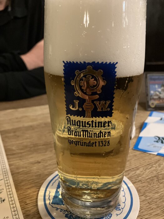 Ett glas öl med skum på toppen, uppställd på ett bord. Glaset har Augustiner Bräu Münchens logotyp.