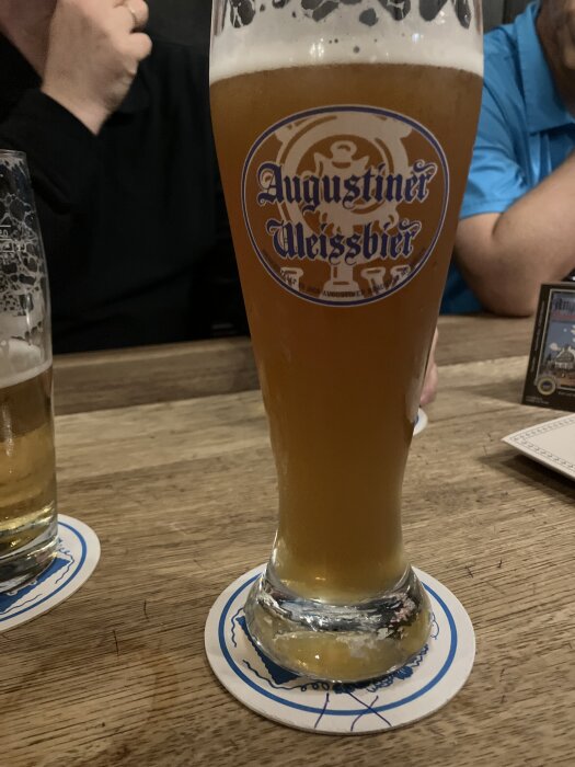 Ett glas Augustiner Weissbier på ett bord med underlägg, omgivet av människor på en restaurang eller pub.