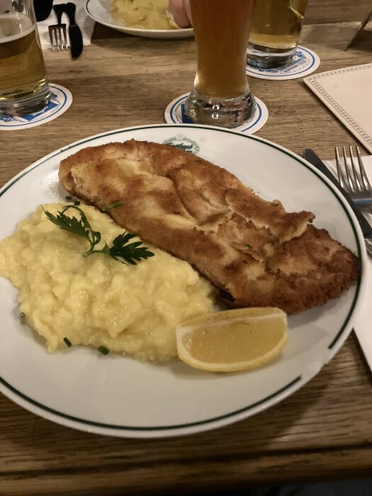 Schnitzel med potatismos, garnerat med en citronklyfta och persilja, serveras på vit tallrik. Ölglas i bakgrunden.