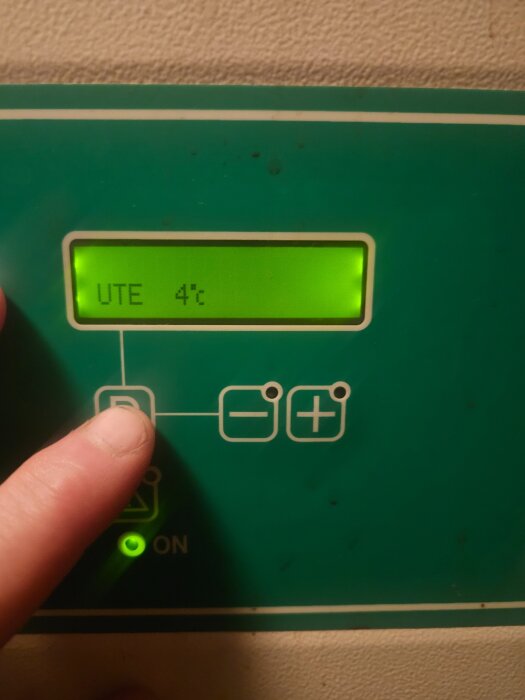 Display på värmepump visar "UTE 4°C", finger pekar på knapp, grön på-lampa lyser.