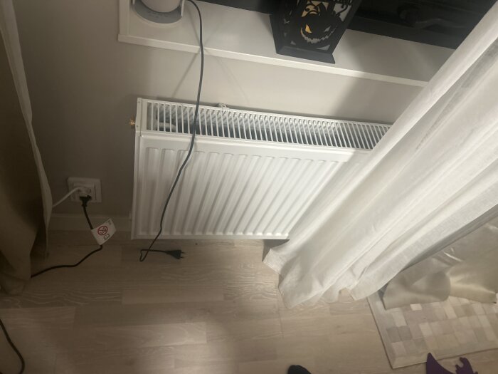 Vitt element under fönsterbräda med sladd och kontakt på ljus trägolv. Vit gardin hänger bredvid; dekor på fönsterbrädan.