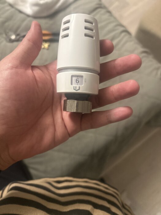 Hand som håller en vit termostatventil i närbild, ett verktyg syns suddigt i bakgrunden.