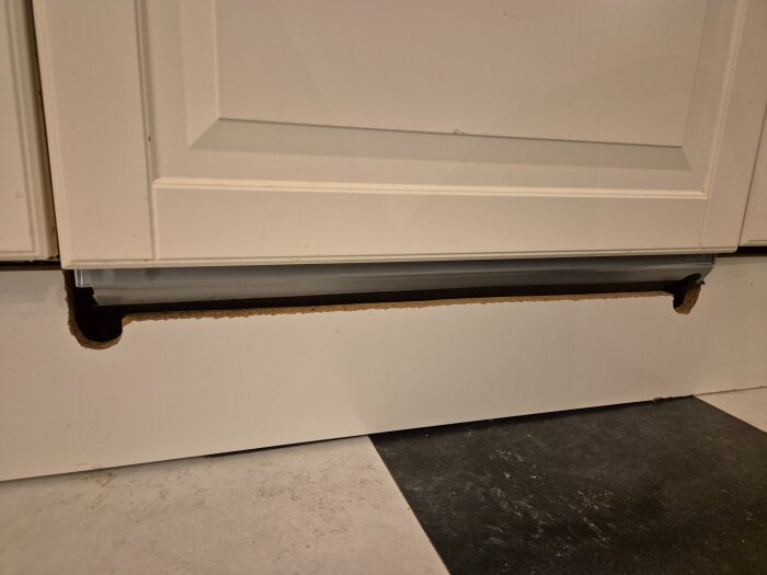 Nedre delen av en köksmaskinfront och en urkapad golvsockel som syns under en vit kökslucka. Golvet är mönstrat i svart och vitt.