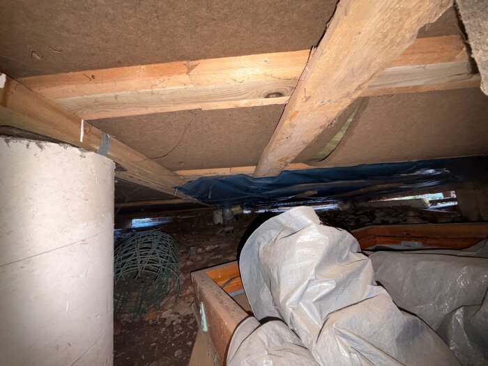Under huset med träbjälkar, oordnad isolering och en plasttäckning. En vit cylinderformad pelare och ett metallnät syns i bakgrunden.