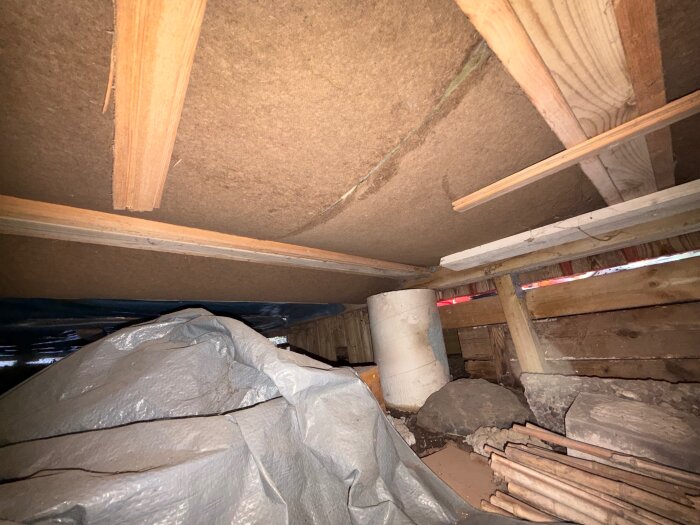 Undersidan av en tillbyggnad med synliga träreglar och isolering, plasttäckt material, och ojämna stenar; potentiella fuktproblem.