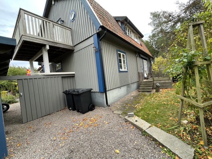 Grått hus med olikåldrad färg, synligt under fönstren. Vädret är mulet. Trädäck och trädgård med gröna växter samt svarta soptunnor.