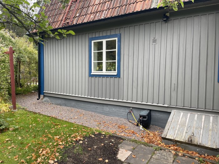 Gråmålad husvägg under fönster i grått väder, visar skillnad i färg mellan nymålat och äldre, samt omgivande gräsmatta och träd.