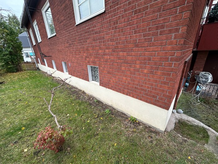 Fasad av tegelhus med grön gräsmatta, buskar och synliga fönsterkällare, potentiellt område för dränering.