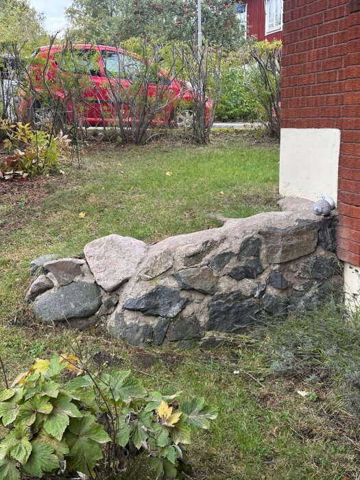 Låg mur av stora stenar vid sidan av ett rött tegelhus, med gräsmatta och buskar i förgrunden.