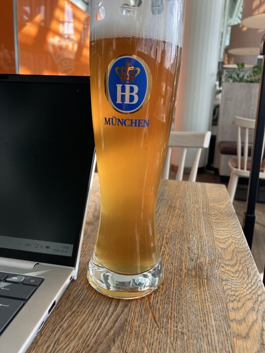 Högt glas med välkomstskum innehållande öl märkt med HB München-logga på ett träbord bredvid en laptop.
