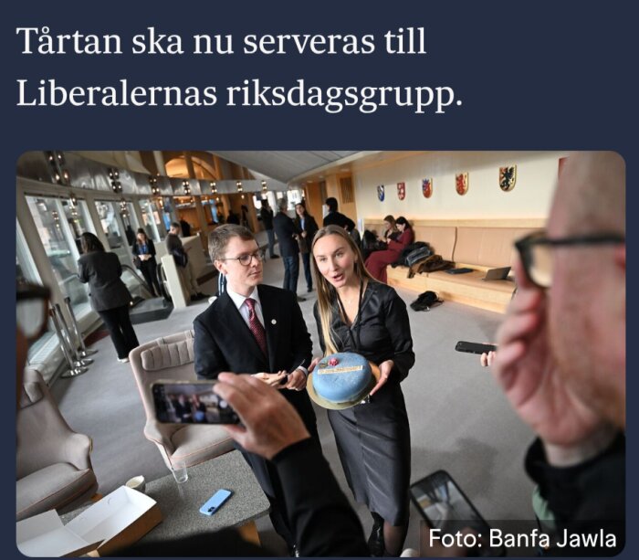 Kvinna håller en tårta i en lokal med flera personer, tårtan ska serveras till Liberalernas riksdagsgrupp.