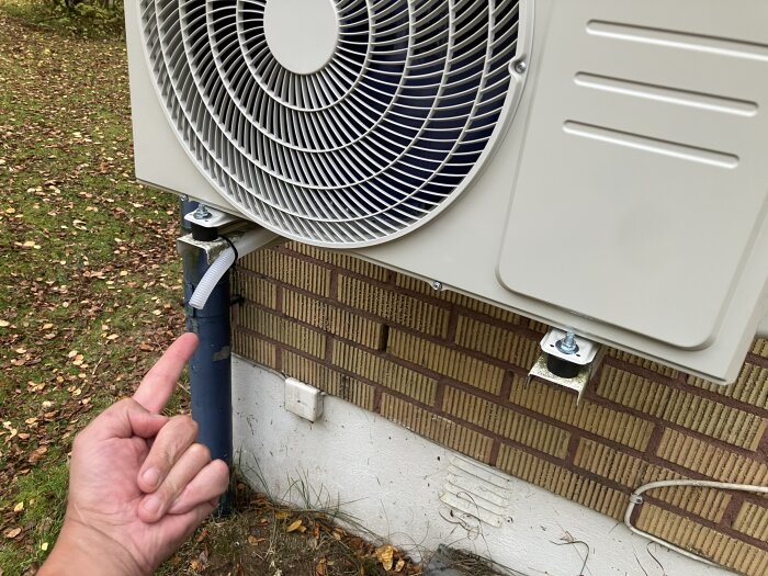 Nyinstallerad värmepump utomhus med avvattningsrör av plast. En hand pekar mot röret mot en bakgrund av tegelvägg och höstlöv.