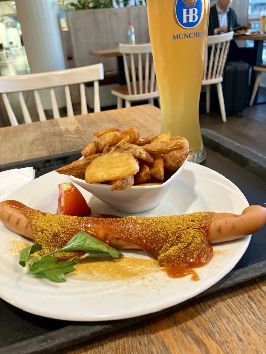 Currywurst med kryddor serverad med potatis och ett glas öl i bakgrunden på ett träbord i en restaurang.