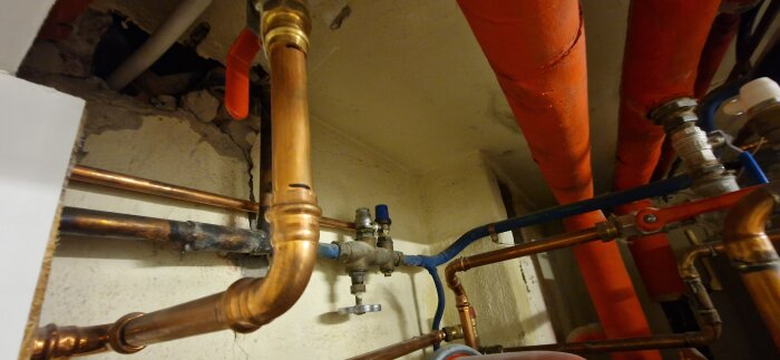 Kopparrör och ventiler i ett trångt utrymme för VVS-installation, med röd och blå isolering på vissa rör.