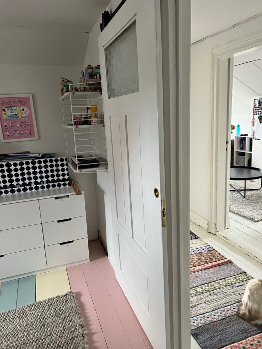 Vit trädörr på glidskena, flerfärgat trägolv och randig matta i ett rum med hyllor, lådor och en hunds bakdel synlig.