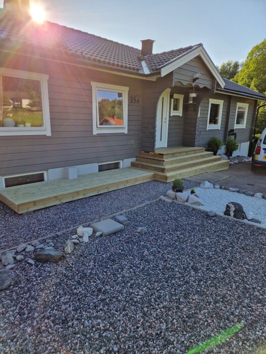 Grått hus med trätrappa och grusad uppfart, solsken i bakgrunden.